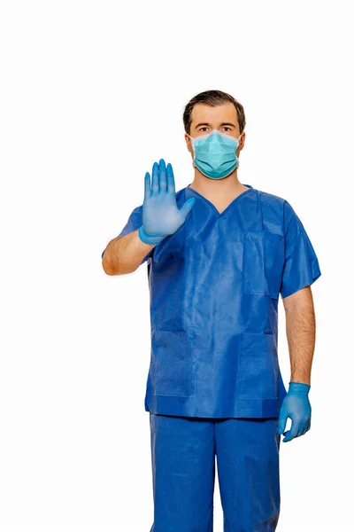 Male doctor in scrubs with protective face mask and gloves showing stop sign with his hand and palm. Plain white background. Coronavirus concept