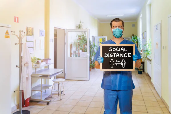 Male doctor in scrubs with protective face mask, holding blackboard with chalk handwriting Social Distance on defocused hospital corridor background