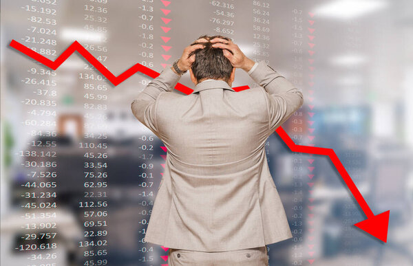 Back view of middle age businessman pulling his hair out as his business falling during recession, data chart and blurred office in background