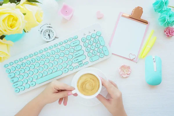 Kadın eller bir fincan kahve, bir bilgisayar faresi ve bir nane beyazı klavye, pembe bir klavye, ataç, bir mum, bir çalar saat, sarı güller tutuyor. Düz düzen, üst görünüm.