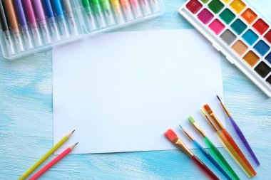 Suluboya resimler, keçeli kalemler, fırçalar, kalemler ve beyaz ve mavi ahşap arka planda beyaz bir kağıt.