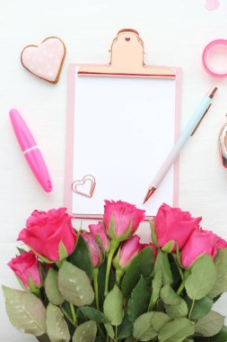 Beyaz bir kağıt, ataç, beyaz bir masa üzerinde pembe ve mavi bir kalem, zencefilli bir kalp ve bir buket parlak pembe gül. Düz düzen, üst görünüm