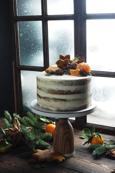 A tall cake on a stand near the glowing window on the windowsill, tangerines, cookies and fir branches