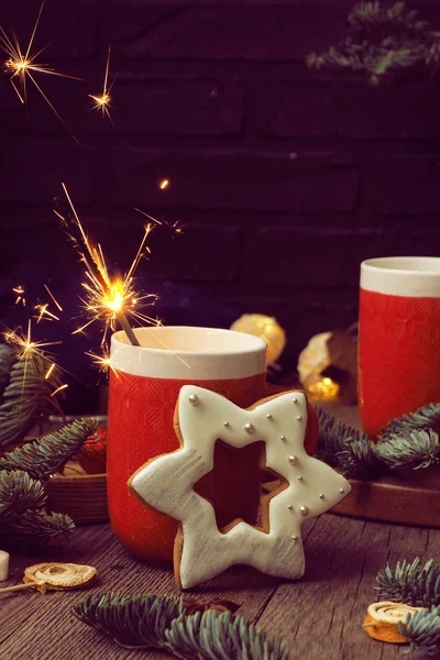 Kahveli ve şekerli iki kırmızı kupa, yanan bir maytap, yıldız şeklinde zencefilli ekmek ve ahşap bir arka planda köknar dalları.