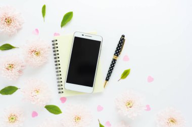 Sarı defterinde siyah ekranı olan beyaz akıllı telefon, puantiyeli siyah kalem, pembe plastik kalpler ve beyaz bir masada yeşil yapraklar. Metin için boşluk