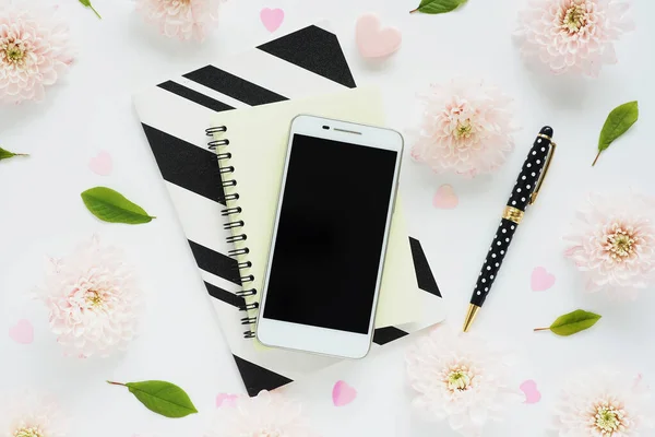 Sarı, siyah ve beyaz çizgili defterleri olan beyaz akıllı telefon üzerinde benekli siyah bir kalem ve beyaz bir masanın üzerinde yeşil yapraklar ve kasımpatı çiçekleri var.