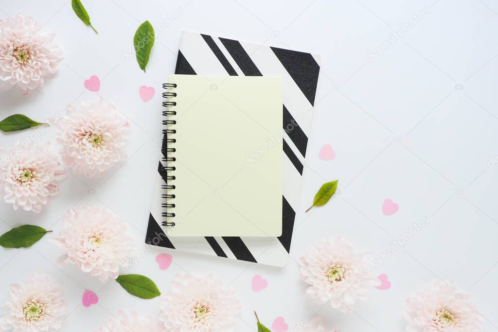 yellow and black and white notebooks, pink plastic hearts and many pink flowers of chrysanthemums and green leaves on a white table. space for text