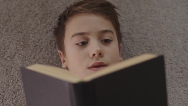 Blick von oben auf einen Jungen, der auf dem Teppichboden ein Buch liest. — Stockvideo