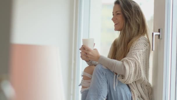 Біла молода жінка п'є чай і дивиться через вікно свого нового будинку. 4k, повільний рух, кишеньковий . — стокове відео
