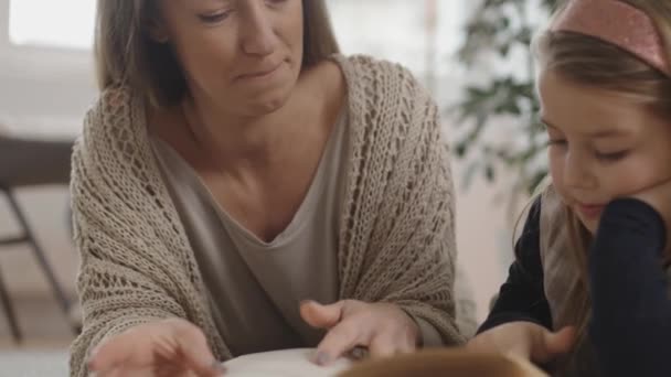 Mamma är stolt över sin dotter medan hon läser en bok på mattan golvet i deras lägenhet — Stockvideo