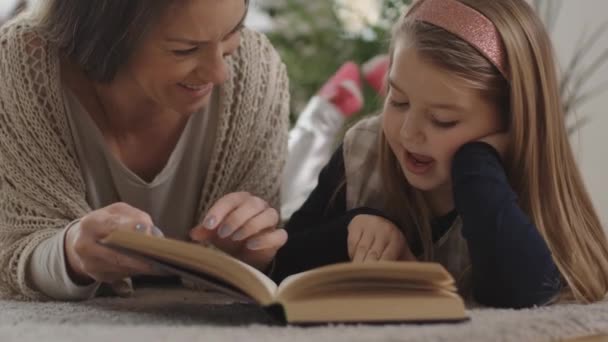 Kleine Tochter und ihre Mutter lesen gemeinsam ein Buch auf dem Teppichboden. — Stockvideo