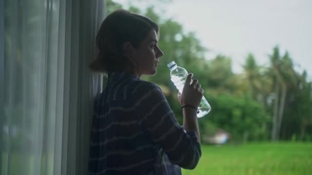 Belle femme caucasienne boire de l'eau en bouteille à son porche avec une belle vue sur la nature vivante . — Video