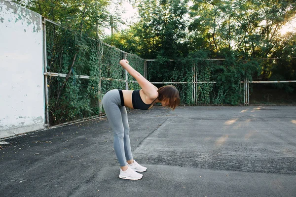 Una chica guapa joven en el patio de recreo en ropa deportiva hace ejercicios físicos. en el verano al atardecer —  Fotos de Stock