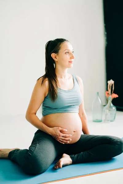 Hermosa chica embarazada en ropa deportiva sentado en una estera de yoga y acariciando su vientre en un estudio — Foto de Stock