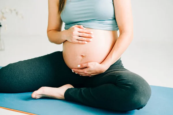 Bela menina grávida em sportswear sentado em um tapete de ioga e acariciando sua barriga em um estúdio — Fotografia de Stock