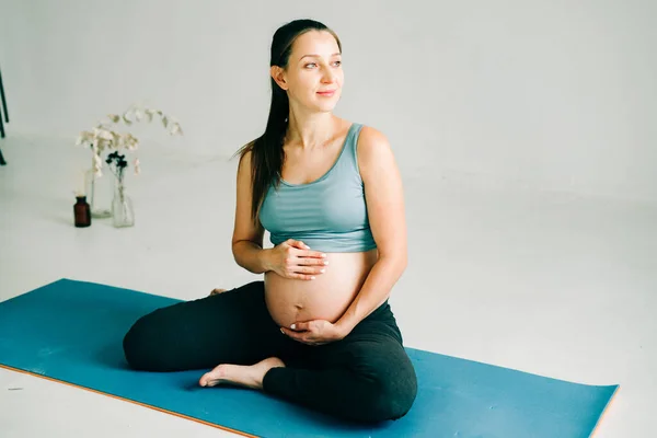 Lodret skud af flot sund og aktiv gravid kvinde i sports bh og leggings, holder håndfladerne sammen og sidder i yoga på rullemåtte derhjemme, mediterer, bliver klar til barsel Stock-foto ©