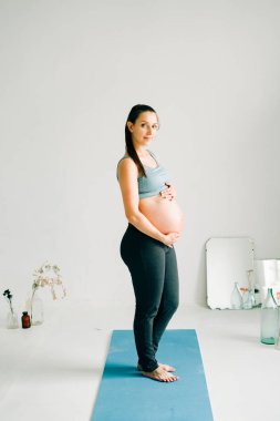 Spor kıyafetli genç hamile bir kız yoga minderinde duruyor, gülümsüyor ve göbeğini stüdyoda tutuyor.