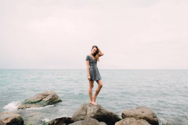 a young girl in a dress stands on the beach on large stones, the wind develops her hair