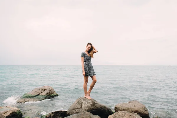 Молода дівчина в сукні стоїть на пляжі на великих каменях, вітер розвиває волосся — стокове фото