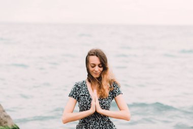 a young beautiful girl in a dress is sitting on large rocks near the sea, meditating and the wind blows her hair
