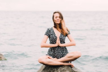 a young beautiful girl in a dress is sitting on large rocks near the sea, meditating and the wind blows her hair