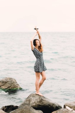 a young girl in a dress stands on the beach on large stones, the wind develops her hair
