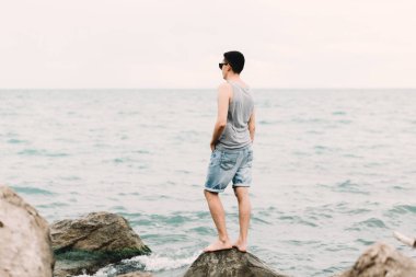 a young guy in a t-shirt and shorts stands on the beach on large rocks