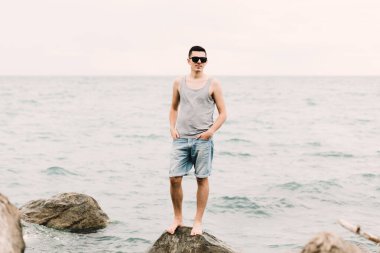 a young guy in a t-shirt and shorts stands on the beach on large rocks