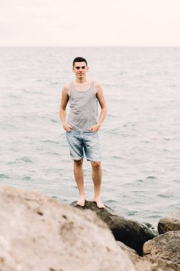 a young guy in a t-shirt and shorts stands on the beach on large rocks