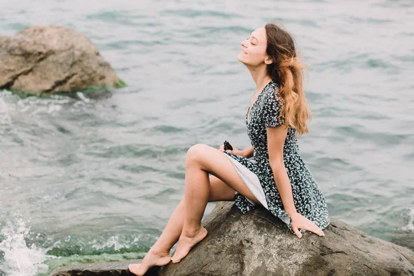 A young beautiful girl in a dress is sitting on large rocks near the sea, the wind is developing her hair — Stock Fotó