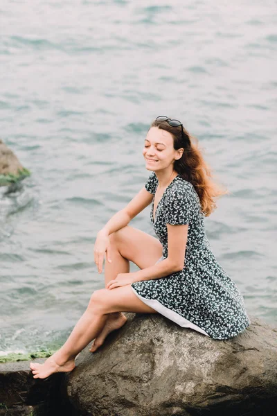 Молода красива дівчина в сукні сидить на великих скелях біля моря, вітер розвиває волосся — стокове фото