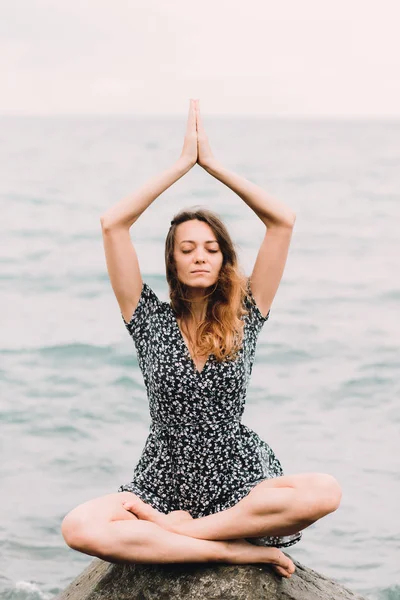 A young beautiful girl in a dress is sitting on large rocks near the sea, meditating and the wind blows her hair — Stock Fotó