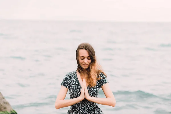 a young beautiful girl in a dress is sitting on large rocks near the sea, meditating and the wind blows her hair