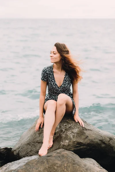 Молода красива дівчина в сукні сидить на великих скелях біля моря, вітер розвиває волосся — стокове фото
