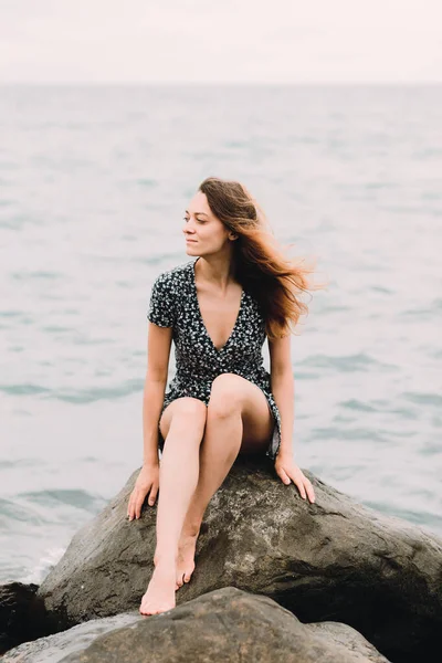 A young beautiful girl in a dress is sitting on large rocks near the sea, the wind is developing her hair — Stock Fotó