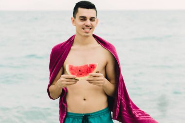 a young guy is standing near the sea with a pink towel on his shoulders and holding a slice of ripe watermelon