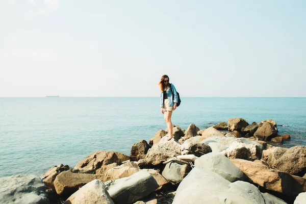 A young girl traveler with a backpack walks barefoot on large stones on the sea coast — Stok fotoğraf