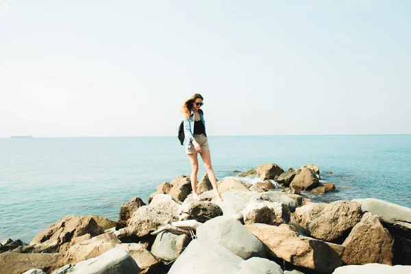 A young girl traveler with a backpack walks barefoot on large stones on the sea coast — Stok fotoğraf