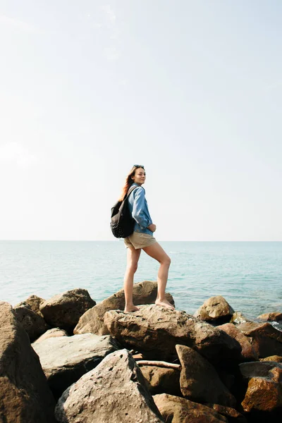 A young girl traveler with a backpack walks barefoot on large stones on the sea coast — Stok fotoğraf