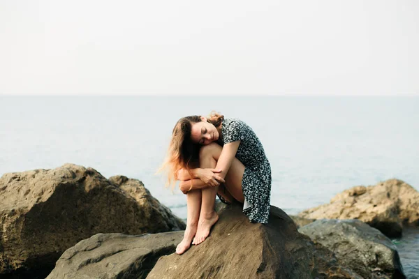 A young beautiful girl in a dress on large stones, a picturesque place on the sea coast — Stok fotoğraf