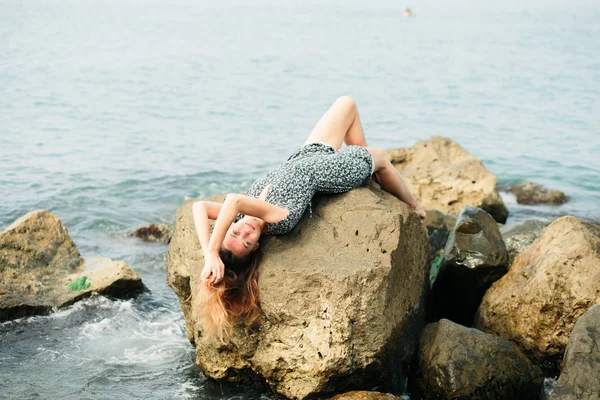 A young beautiful girl in a dress on large stones, a picturesque place on the sea coast — Stok fotoğraf