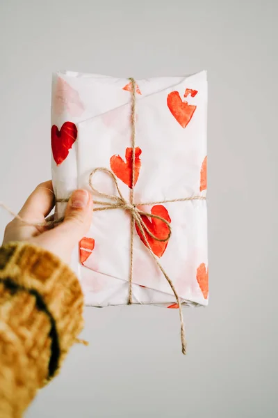 Um presente na mão de uma mulher envolto em papel de embrulho caseiro com corações vermelhos amarrados com fio de juta para o dia dos namorados — Fotografia de Stock