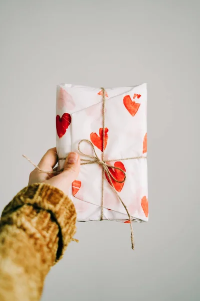 A gift in a woman's hand wrapped in homemade wrapping paper with red hearts tied with jute thread for Valentine's day — Stok fotoğraf