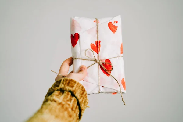 Um presente na mão de uma mulher envolto em papel de embrulho caseiro com corações vermelhos amarrados com fio de juta para o dia dos namorados — Fotografia de Stock