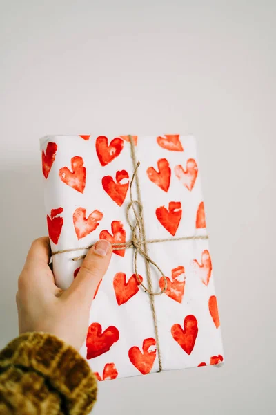 A gift in a woman's hand wrapped in homemade wrapping paper with red hearts tied with jute thread for Valentine's day — Stock Photo, Image