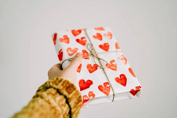 Um presente na mão de uma mulher envolto em papel de embrulho caseiro com corações vermelhos amarrados com fio de juta para o dia dos namorados — Fotografia de Stock
