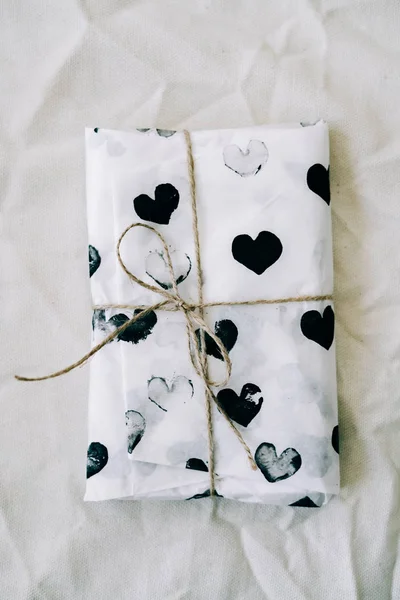 Um presente embrulhado em papel de embrulho caseiro com corações pretos amarrados com fio de juta para o dia dos namorados em uma mesa branca — Fotografia de Stock
