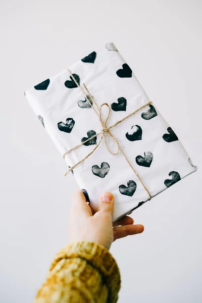 Um presente bonito na mão de uma mulher envolto em papel de embrulho caseiro com corações pretos amarrados com fio de juta para o dia dos namorados em um fundo branco — Fotografia de Stock