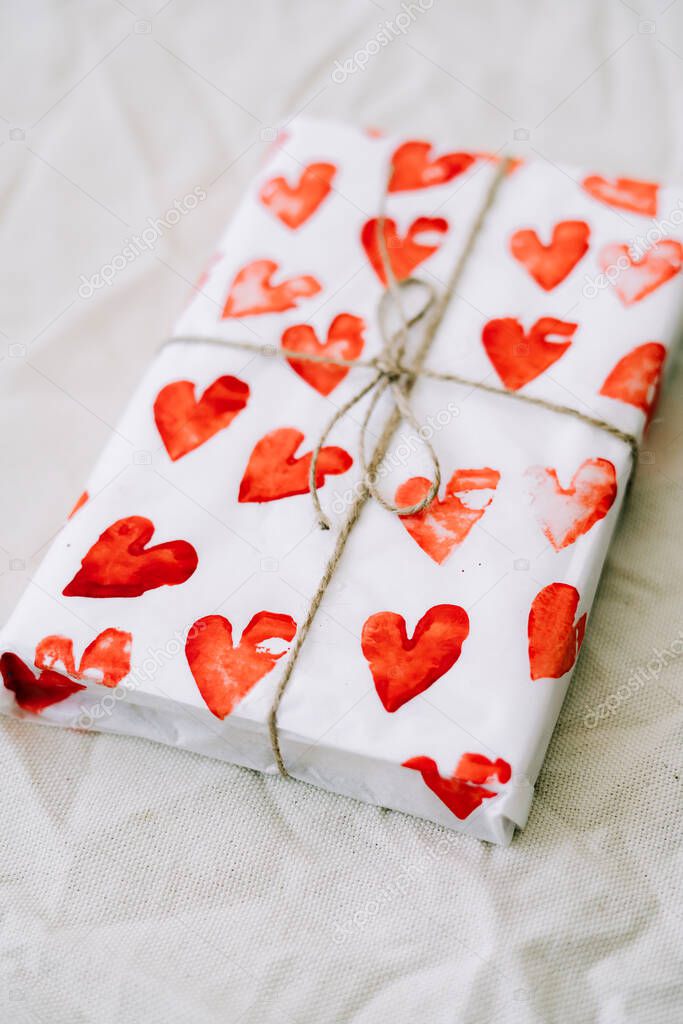 a gift wrapped in homemade wrapping paper with red hearts tied with jute thread for Valentine's day on a white table