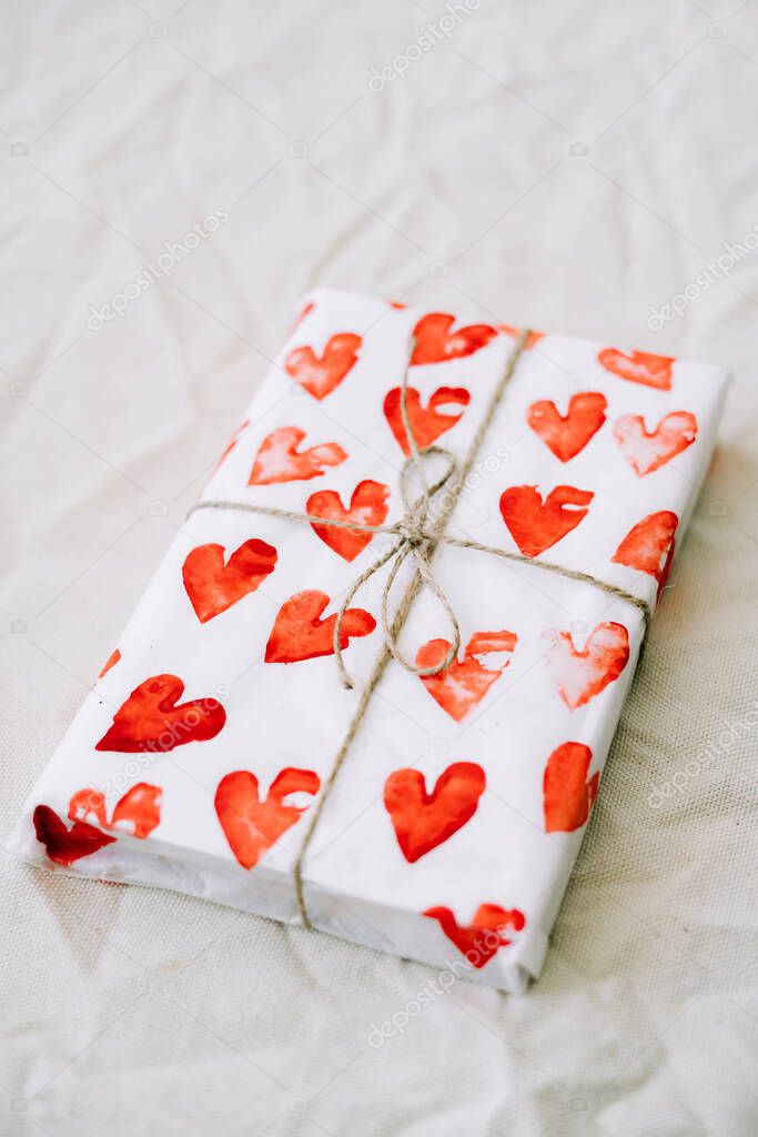 a gift wrapped in homemade wrapping paper with red hearts tied with jute thread for Valentine's day on a white table